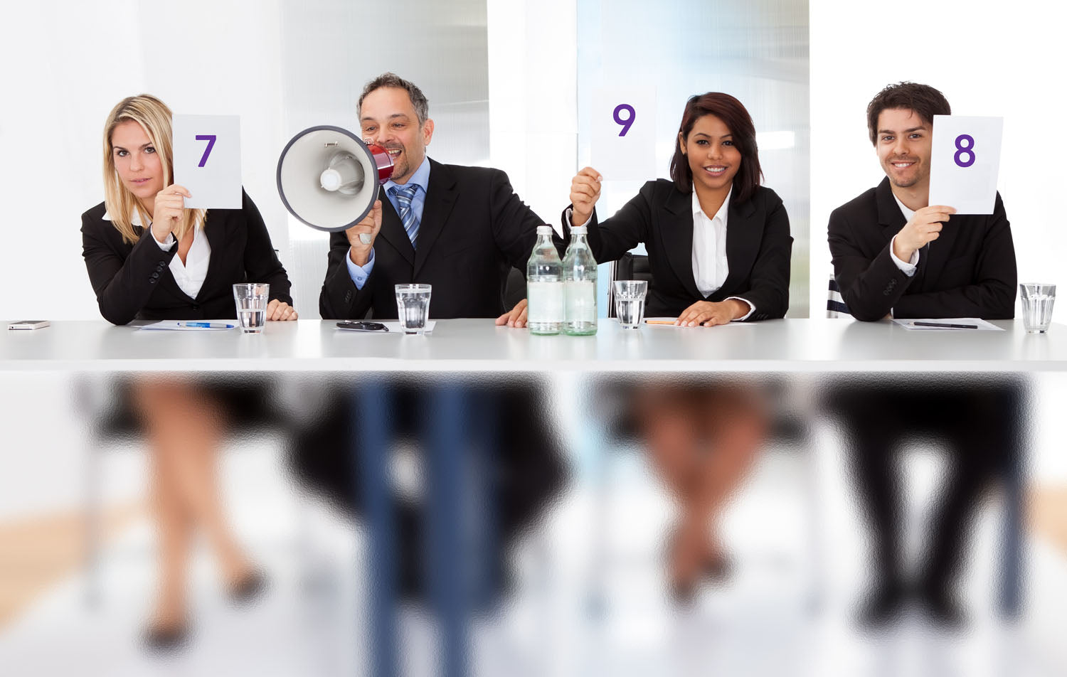 Four people in business suits behind a table.  Three hold score placards reading 7, 9, 8.  The person 2nd from the left holds a megaphone (licensed from Adobe Stock Photos)