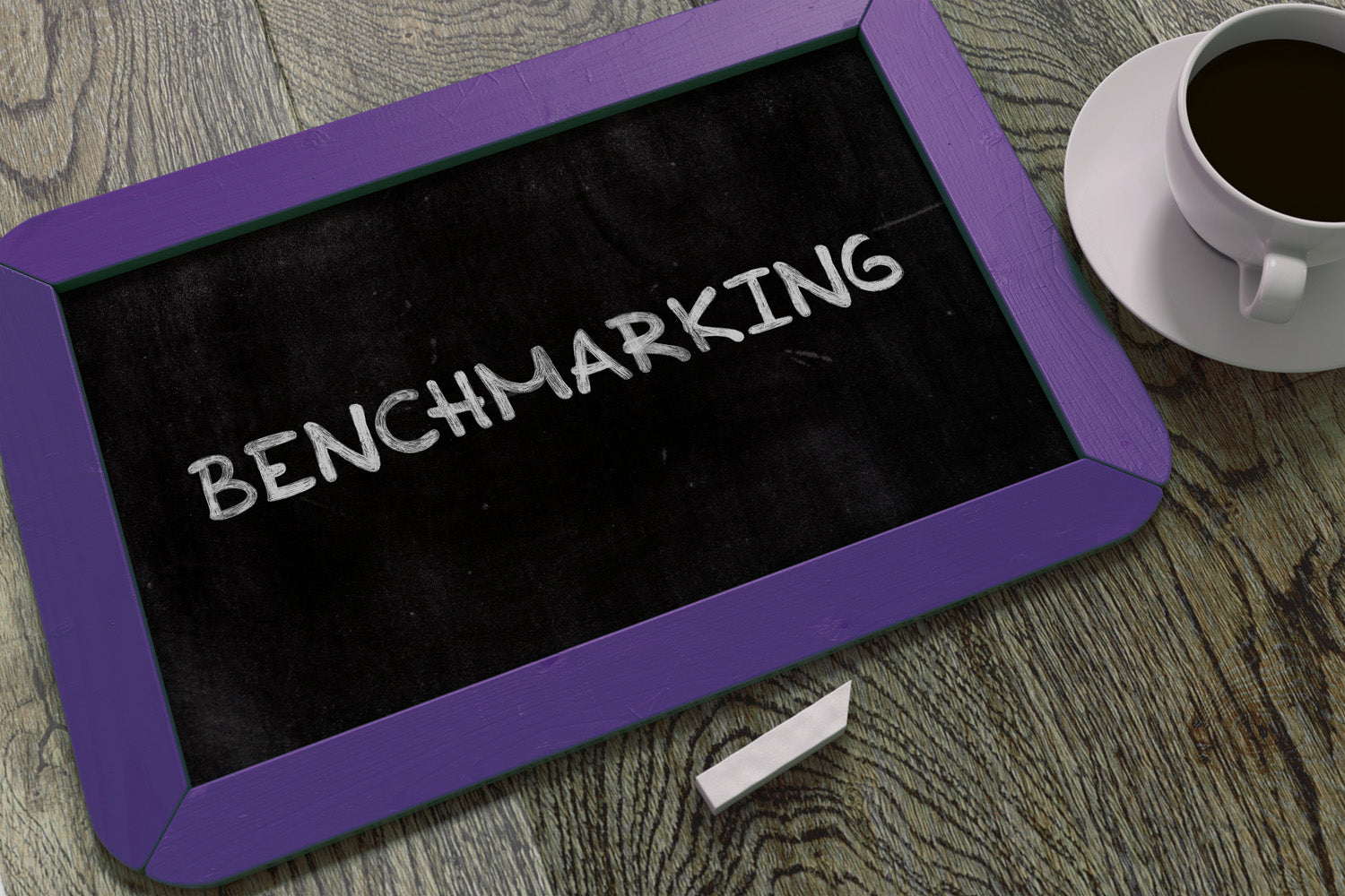 Benchmarking written on a small tablet-sized blackboard sitting on a table next to a cup of coffee (licensed from Adobe Stock Photos)
