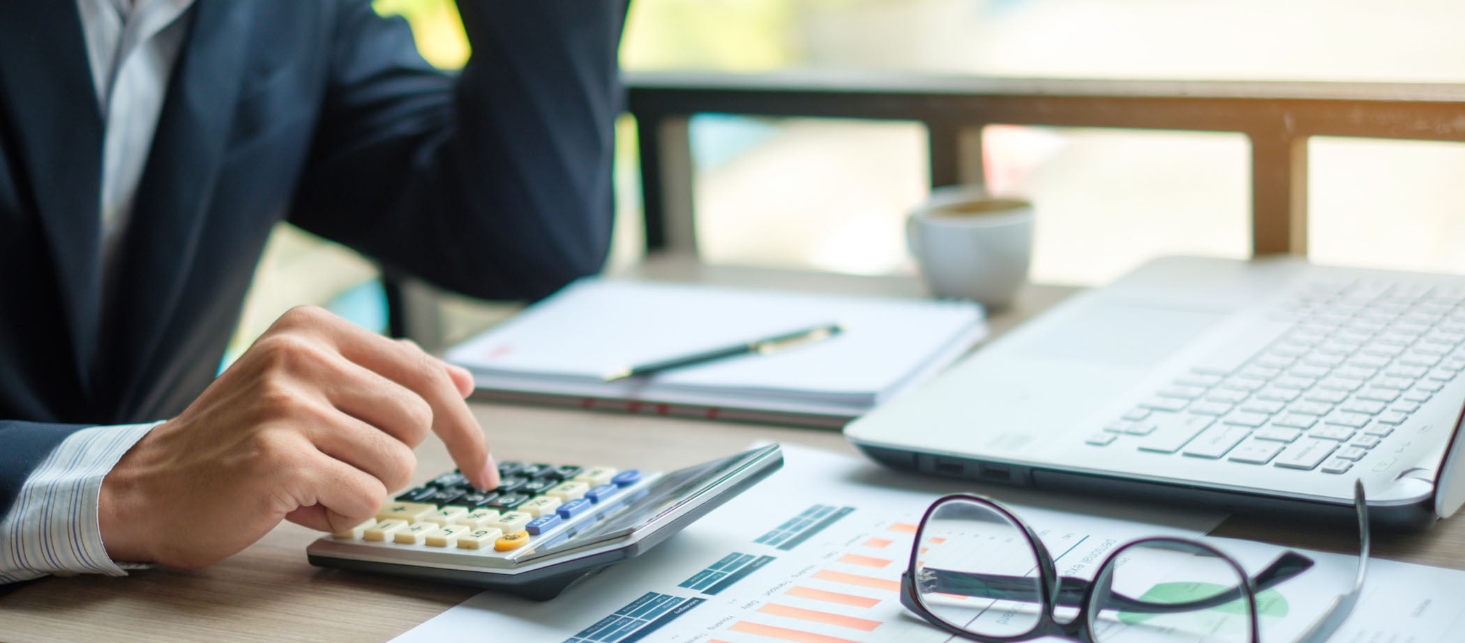 close-up of business person in suit tapping calculator with a laptop graphs on paper and reading glasses lying on the table (licensed from Adobe Stock Photos)